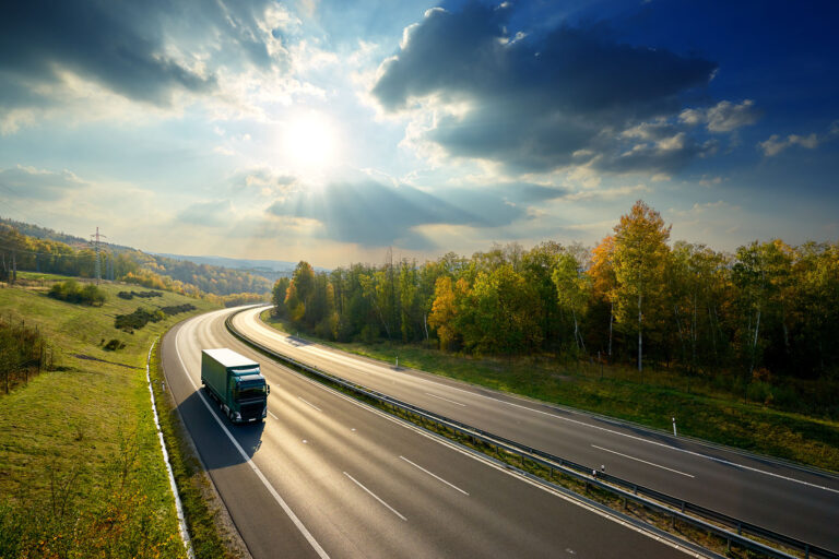 Green truck driving on the asphalt highway between deciduous for